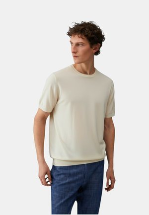 Giovane con capelli corti e ricci, indossa una maglietta crema a maniche corte tinta unita e pantaloni blu a quadri, guarda verso sinistra.