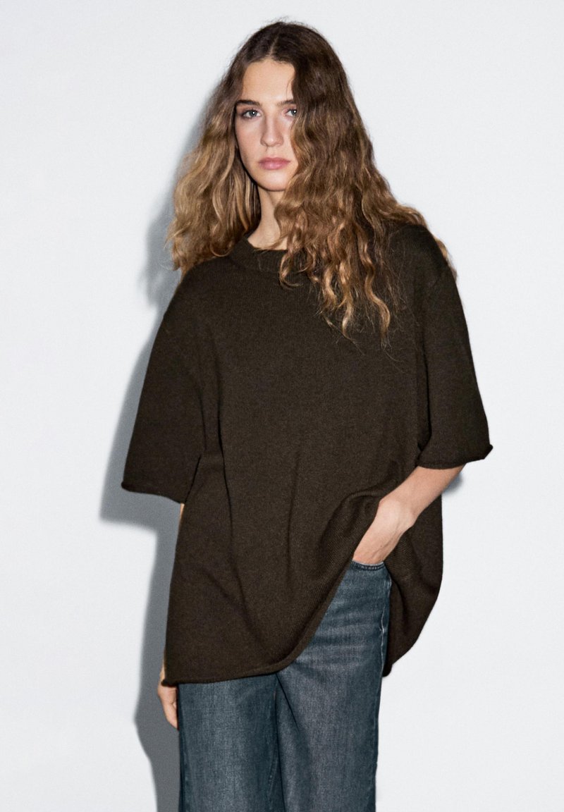 Maglione oversized marrone scuro a maglia con maniche corte, scollatura tonda e vestibilità comoda, abbinato a jeans denim blu chiaro.