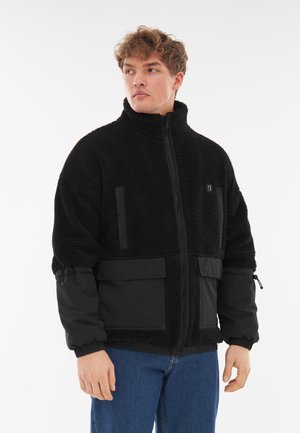 Giacca in fleece nera con colletto alto, chiusura con zip, tasche frontali e polsini elasticizzati. Presenta un tessuto testurizzato e pannelli lisci.