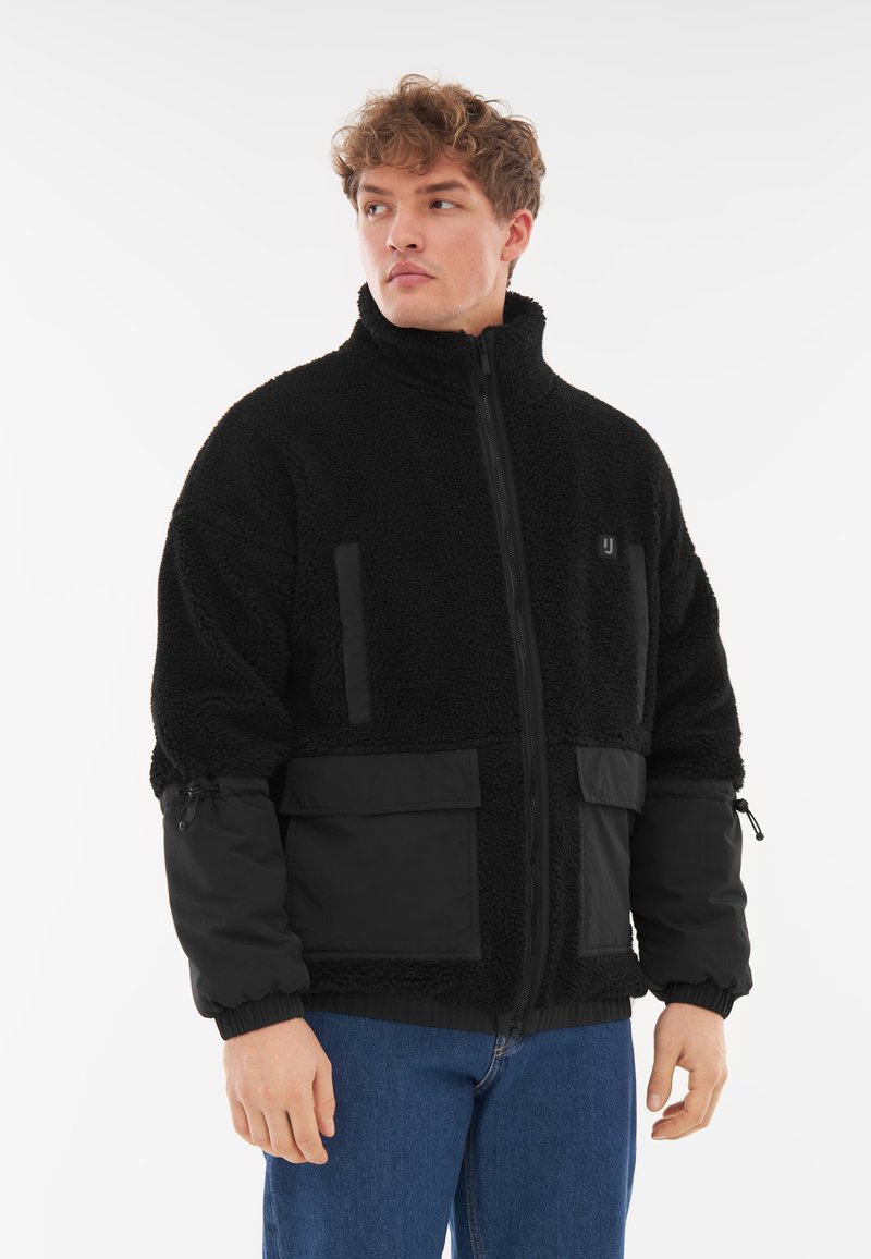 Giacca in fleece nera con colletto alto, chiusura con zip, tasche frontali e polsini elasticizzati. Presenta un tessuto testurizzato e pannelli lisci.