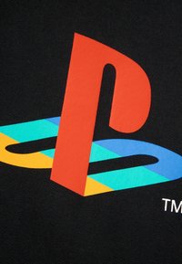 Czerwony znak "P" PlayStation na kolorowym "S" w odcieniach niebieskiego, żółtego i turkusowego na czarnej tkaninie, część logo PlayStation.