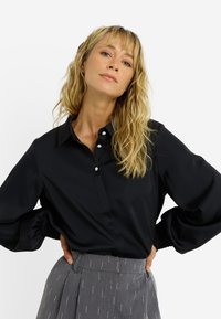 Chemise noire à manches longues en satin avec trois boutons décoratifs, dotée d'un col et de poignets froncés, associée à un pantalon gris à motifs.