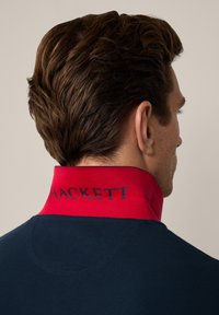 Marineblå polo skjorte med en fremtredende rød krage med ordet "HACKETT" i mørkeblått. Glatt tekstur, klassisk design og tilpasset passform.