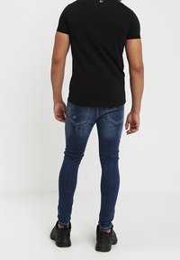 Slim-fit černé tričko, modré džíny s vybledlým umytím, červené prošívání na zadních kapsách, doplněné černými sportovními botami.