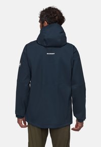 Veste imperméable bleu marine avec une capuche, présentant un logo au dos, fabriquée en matériau léger et lisse avec des poignets élastiques.