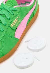Baskets Puma Palermo en daim vert avec une bande rose et une semelle en gomme, accompagnées de deux lacets blancs enroulés sur un fond blanc.