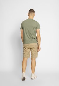 T-shirt verde oliva semplice abbinato a pantaloni corti beige. La maglietta è a maniche corte e i pantaloni corti hanno tasche laterali. Scarpe alte beige.