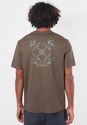 Personne aux cheveux bouclés porte un t-shirt marron avec un motif floral bleu et vert et le texte "OXBOW" au dos.