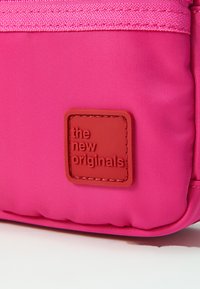 Borsa in tessuto rosa con superficie texturizzata, caratterizzata da una toppa logo in rilievo rossa "the new originals" e chiusura con zip nella parte superiore.