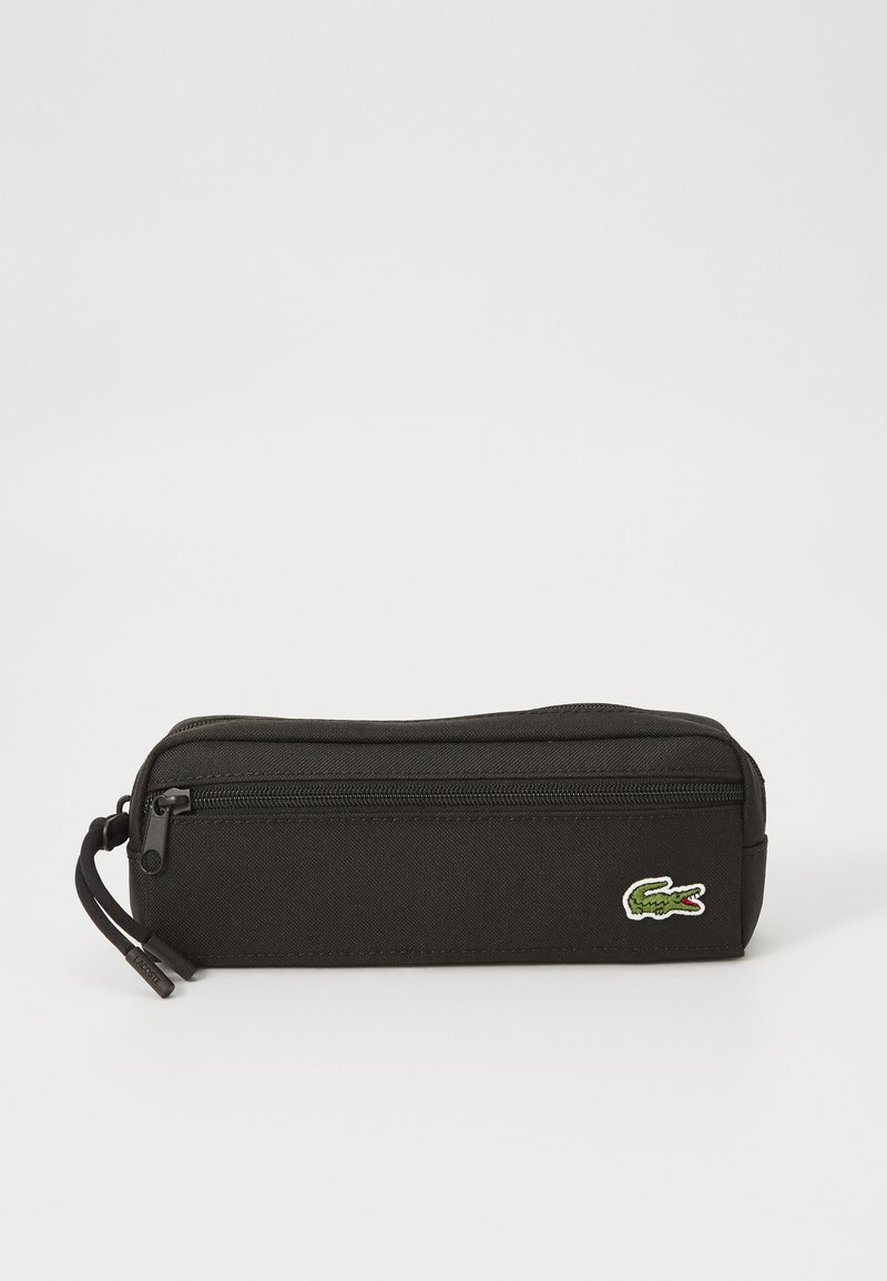 Trousse noire en tissu texturé, avec fermeture éclair et un petit logo de crocodile vert sur le côté. Forme rectangulaire.
