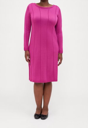 Mujer vestida con un vestido ajustado de manga larga hasta la rodilla, de color rosa brillante, con detalles de costuras verticales y zapatos planos negros.