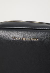 Juodų odinių krepšys su tekstūruotu paviršiumi, auksinės spalvos logotipas "TOMMY HILFIGER" ir užtrauktuku uždaromas viršus.