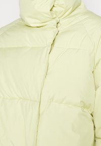 Doudoune jaune clair avec col montant, design matelassé et coutures diagonales sur la poitrine et les épaules.