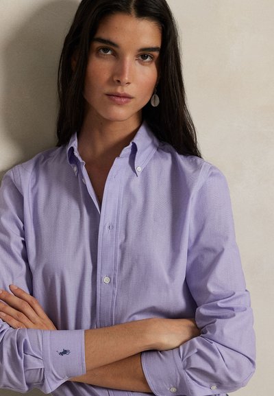 Pitkät tummahiuksinen nainen, jolla on päällään vaaleanvioletti nappipaita, seisoo käsivarret ristissä neutraalin taustan edessä.