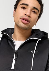 Svart vindjacka med en vit dragkedja, med en Nike-logotyp på bröstet och vita snoddar på huvan. Slät textur.