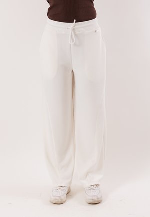 Persona che indossa pantaloni bianchi a gamba ampia con vita con coulisse e sneakers bianche, in piedi contro uno sfondo chiaro uniforme.