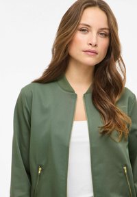 Jachetă bombero verde cu textură netedă, guler striat, fermoar auriu și buzunare laterale, purtată peste un top alb.