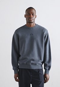 Sweatshirt cinza em tecido suave com decote redondo e punhos canelados. Apresenta um logotipo discreto ao centro. Usado com calças escuras.