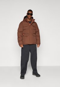 Braune Steppjacke mit Kapuze, Reißverschluss vorne, Brust- und Seitentaschen, kombiniert mit schwarzen Hosen und robusten schwarzen Schuhen.