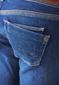 Modré džíny z denimu mají zadní kapsu s kontrastním šitým vlnitým vzorem, hnědou koženou nášivkou a kovovým knoflíkem.