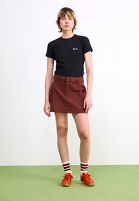 T-shirt in cotone nero, gonna in denim color ruggine con tasche frontali, calzini a strisce e sneaker arancioni su pavimento verde.