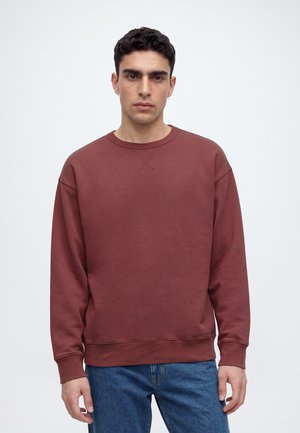 Un homme se tient devant un fond clair et uni, vêtu d’un sweat-shirt bordeaux à col rond et d’un jean bleu.