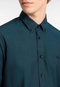 Teal button-up shirt gemaakt van geweven stof, met een klassieke kraag en zwarte knopen. Klein geborduurd logo op de borst.