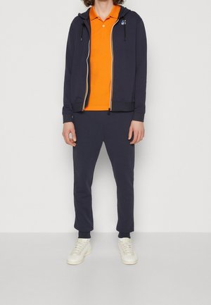 Osoba v tmavě modré mikině na zip, oranžovém polokošili, sladěných tmavě modrých teplácích a bílých teniskách, stojící proti jednoduchému světlému pozadí.