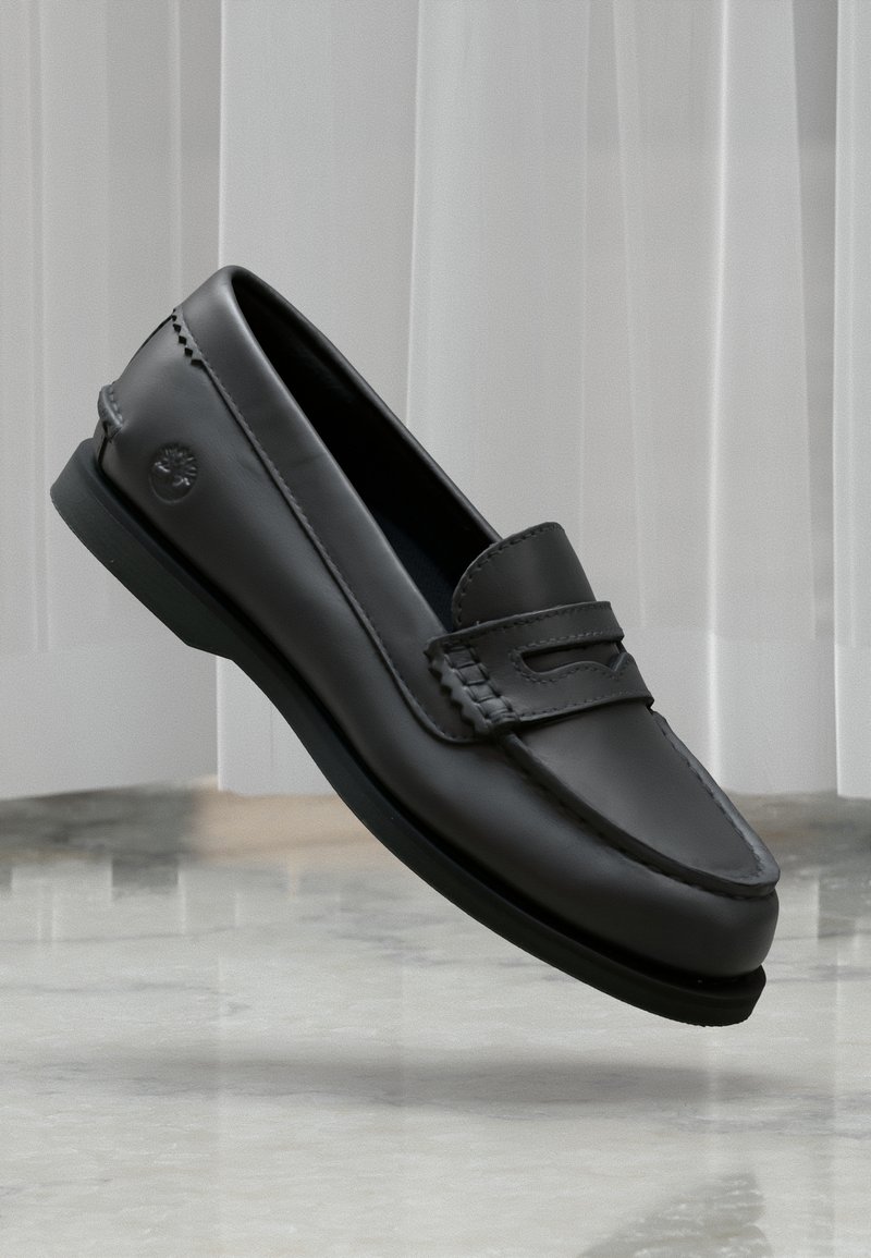 Mocassin en cuir noir avec détails cousus et logo sur le côté, présenté en suspension au-dessus d'un sol en marbre sur un fond de rideau clair.