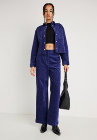 Γυναίκα που φοράει μπλε κοτλέ ζακέτα και παντελόνι με μαύρη κοντή μπλούζα, κρατώντας μια μαύρη τσάντα και φοράει μαύρες μυτερές μπότες.