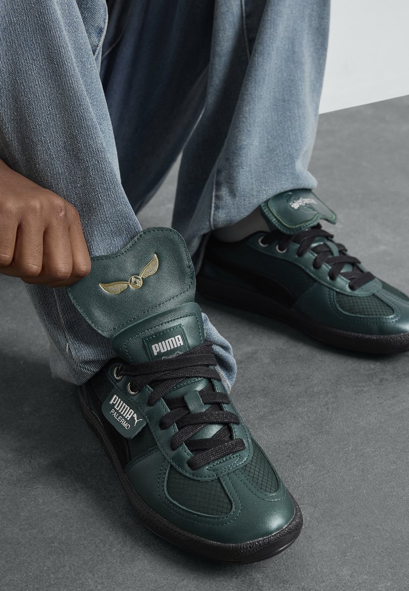 Main remontant la jambe du pantalon pour révéler des baskets en cuir vert foncé Puma Palermo avec des lacets noirs et un emblème ailé doré sur la languette.