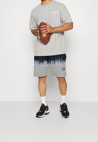 T-shirt grigio a righe, pantaloni corti neri e grigi con effetto tie-dye, scarpe da ginnastica nere, tiene un pallone di football marrone. Abbigliamento sportivo semplice e informale.