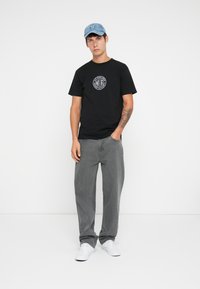 Black cotton t-shirt with circular white logo, paired with gray loose-fit jeans and white sneakers. Light blue cap with embroidered detail.