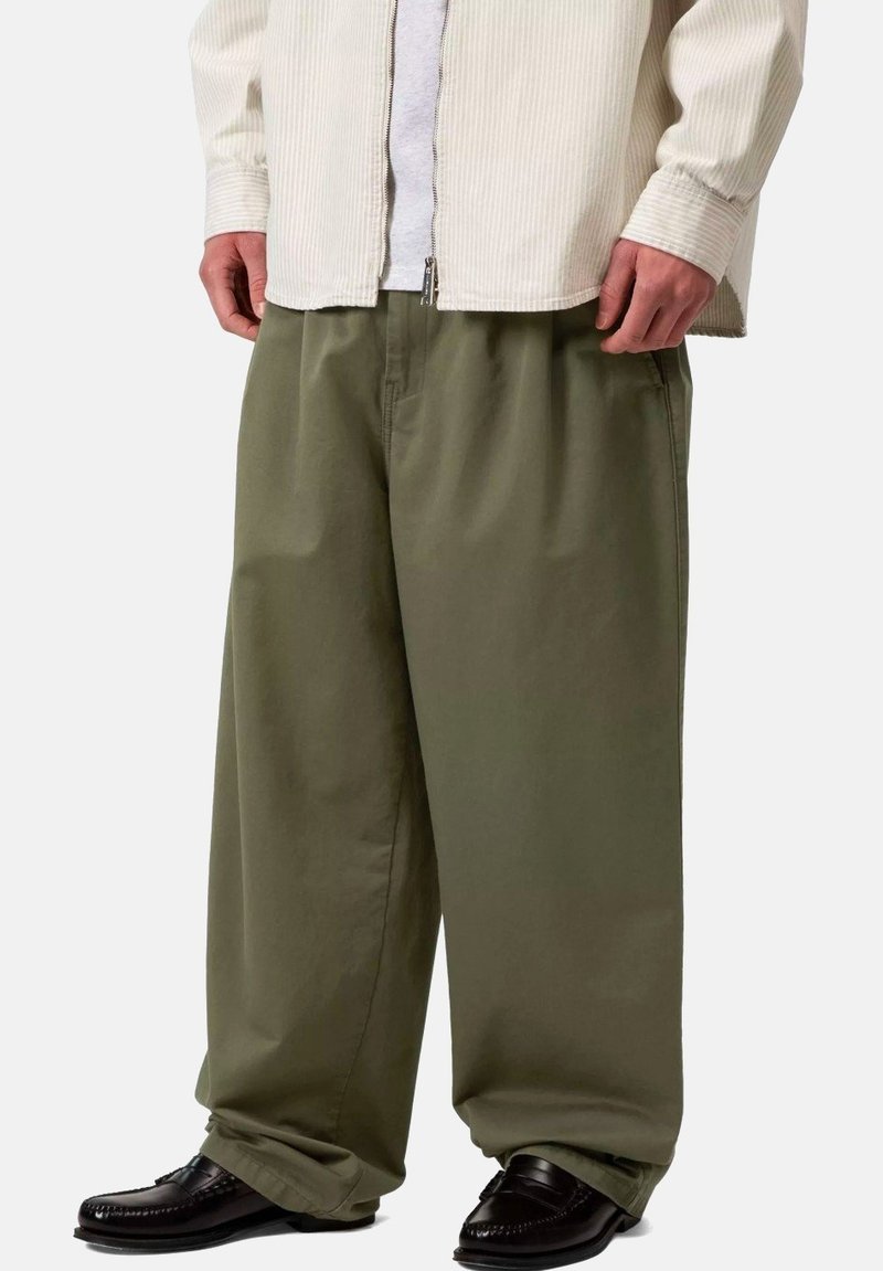 Hombre con pantalones anchos de color verde oliva, zapatos de cuero negros y una camisa de pana beige sobre una camiseta blanca, de pie contra un fondo liso.