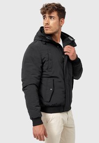 Black padded jacket with a hood, front zip, two side pockets, ribbed cuffs, and a logo detail on the collar. Smooth texture.