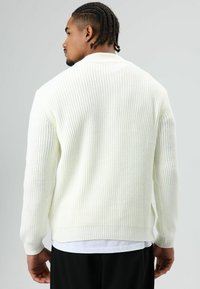 Man with braided hair wearing a white ribbed sweater, white undershirt, and black pants, facing away against a plain grey background.