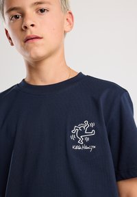 Navy cotton t-shirt featuring a white Keith Haring graphic on the left chest. Classic crew neck design and short sleeves.