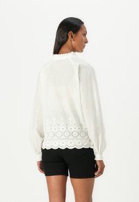 Hvit bluse med brodert blomstermønster og scallop-kant, med lange ermer og volang-halskant, brukt med sorte shorts.