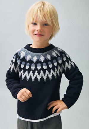 Enfant blond portant un pull bleu marine avec des motifs géométriques blancs et gris, se tenant avec une main sur la hanche et un léger sourire.