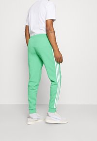 Pantalones de chándal verdes con rayas verticales blancas en los lados. Están hechos de un material suave, presentan un ajuste ajustado y una cinturilla elástica.