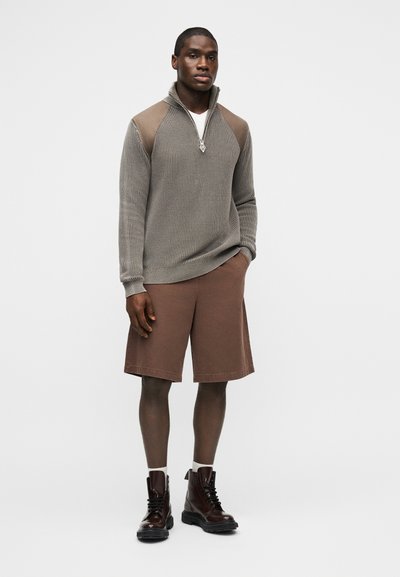 Άντρας που φοράει γκρι πουλόβερ με φερμουάρ μέχρι τη μέση και ταμπά επιθέματα στους ώμους, καφέ σορτς μέχρι το γόνατο, λευκές κάλτσες και σκούρα καφέ μπότες με κορδόνια.