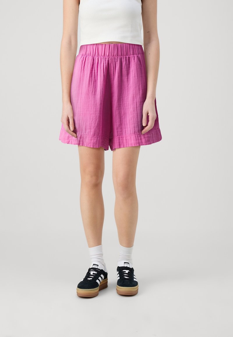 Personne portant un short ample rose vif, un crop top blanc, des chaussettes blanches et des baskets noires, debout devant un fond clair uni.