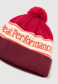Adīta cepure bordo un sarkanā krāsā ar baltu un oranžu joslu, uz kuras ir uzraksts "Peak Performance." Mīksta tekstūra, ar atbilstošu pom-pom virsotnē.