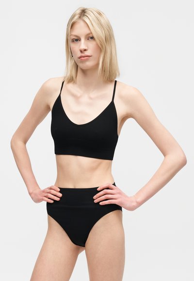Νεαρή γυναίκα με ξανθά μαλλιά φορώντας μαύρο κοντό τοπ και μαύρο εσώρουχο με ψηλή μέση, όρθια με τα χέρια στη μέση.