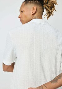 Chemise blanche à manches courtes en tissu texturé avec un motif subtil. Dotée d'un col classique et d'une coupe décontractée, mettant en valeur un tatouage sur le côté.