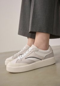 Sneaker bianchi e grigio chiaro con finitura in pelle texture, dotati di punta arrotondata, lacci bianchi e una suola in gomma spessa.