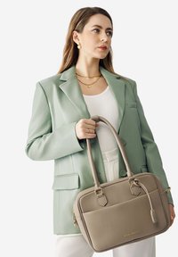 Donna in un blazer verde chiaro che tiene una borsa taupe con texture e dettagli dorati, indossando una maglietta bianca, collane d'oro e orecchini.