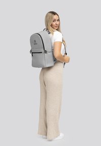Zaino grigio con parte superiore arrotondata, tasca frontale con zip e accenti neri. Il modello indossa una camicia bianca e pantaloni beige, in piedi su uno sfondo neutro.