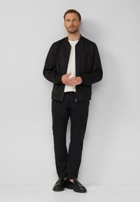 Zwarte bomberjack met ritssluiting, gedragen over een wit T-shirt, gecombineerd met zwarte broek en zwarte schoenen, op een grijze achtergrond.