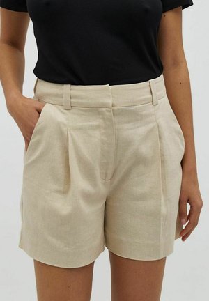 Person trägt beige Leinen-Shorts mit Falten und Gürtelschlaufen, kombiniert mit einem schwarzen kurzärmligen Oberteil, die Hände entspannt an den Seiten und in der Tasche.